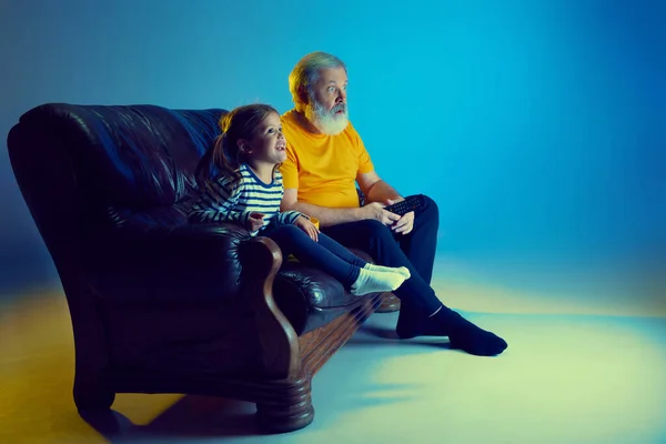 Senior man and little girl, grandfather watching TV with his granddaughter over blue background in neon light. Concept of family, happiness, care and love, unity, emotions, leisure