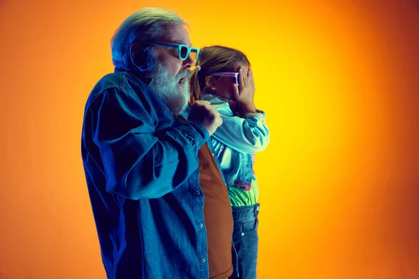 Horror movie. Senior man watching movie with little girl over gradient orange background in neon light. Concept of family, happiness, care and love, unity, emotions, leisure