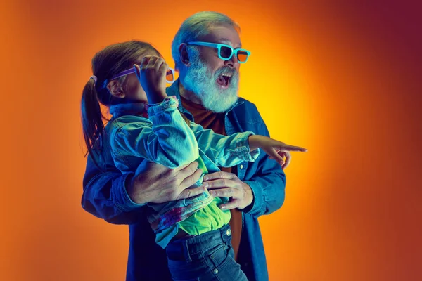 Grandfather watching movie with his little granddaughter over gradient orange background in neon light. Concept of family, happiness, care and love, unity, emotions, leisure