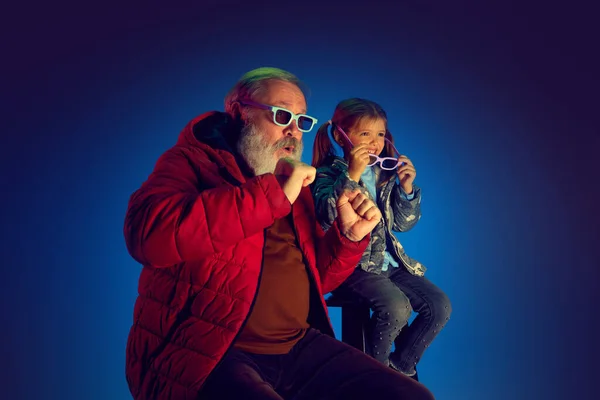 Emotional grandfather and little girl, child sitting and watching TV over blue background in neon light. Concept of family, happiness, care and love, unity, emotions, leisure