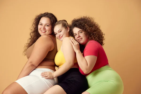 Friends, beautiful women with oversized bodies, wearing sportswear, posing over beige studio background. Concept of sport, body-positivity, weight loss, body and health care