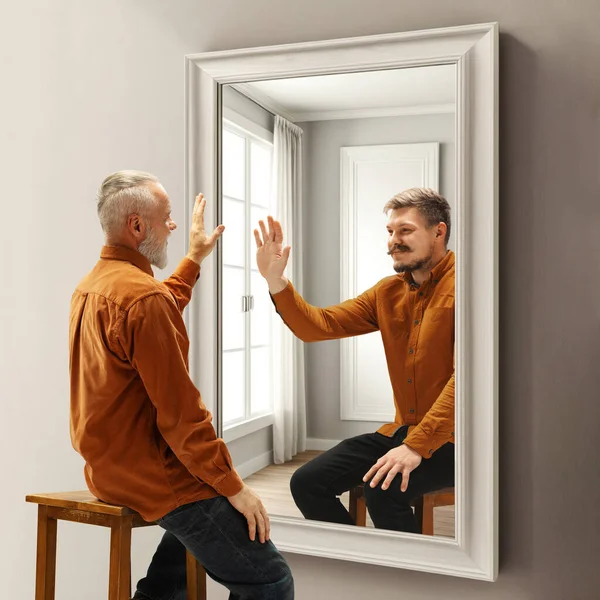 Senior smiling man looking in mirror on reflection of his younger self. Conceptual collage. Past memories. Concept of present, past and future, age, life cycle, generation, ad
