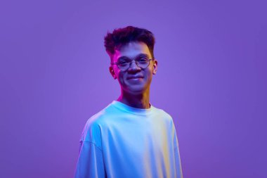 Gülümseyen genç bir adamın portresi, günlük beyaz tişörtlü ve güneş gözlüklü bir çocuk neon ışıkta mor arka planda kameraya bakıyor. İnsan duyguları, gençlik yaşam tarzı, moda kavramı. Reklamlar