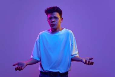 Sıradan tişörtlü ve gözlüklü çocuk ellerini açıyor, yanlış anlama yüzüyle neon ışıkta mor arka planda duruyor. İnsan duyguları, gençlik, yüz ifadesi kavramı