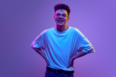Günlük beyaz tişörtlü, kot pantolonlu ve gözlüklü mutlu genç adam gülümsüyor, neon ışıkta mor arka plana gülüyor. Mutluluk, neşe ve eğlence. İnsan duyguları, gençlik, başarı kavramı