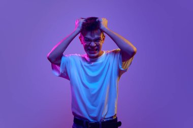 Gündelik giysiler içindeki genç adam kafasının üstünde el ele tutuşuyor, neon ışıkta mor arka plana karşı öfke, sinir ve stresi ifade ediyor. İnsan duyguları, gençlik, psikoloji, iç dünya kavramı