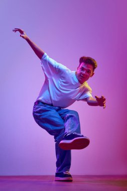 Günlük kıyafetler, kot pantolon ve beyaz tişörtler içinde neon ışıkta hip hop yapan neşeli genç bir adam. Hobi, aktif yaşam tarzı. İnsan duyguları kavramı, gençlik kültürü, moda