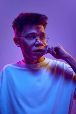 Beyaz tişörtlü ve gözlüklü komik bir gencin portresi komik surat yapan, elleri neon ışıkta mor arka planda uzanan ağzı olan. Meme. İnsani duygular, gençlik, eğlence ve neşe kavramı