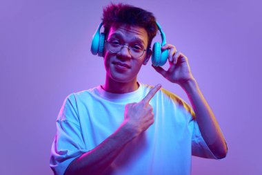 Gözlüklü genç adam kulaklık takıyor, parmaklarıyla ona işaret ediyor neon ışıkta mor arka planda. İyi ses, yüksek kaliteli aksesuar. İnsan duyguları, gençlik, müzik, aletler kavramı