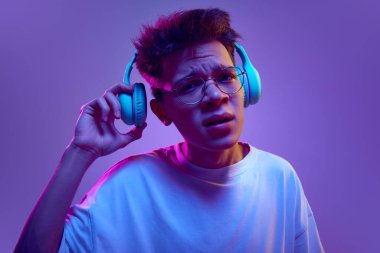 Kulaklıklı sorgulayan, kulaklıklı, kulaklıklı genç bir adam. Mor arka planda, neon ışıkta net bir şekilde duyabilmek için kulaklıklarını çıkarıyor. İnsan duyguları, gençlik, müzik, aletler kavramı