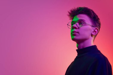 Portrait of young man, student in stylish clothes and glasses standing with proud face against gradient pink background in neon light. Concept of human emotions, youth, growth, achievement clipart