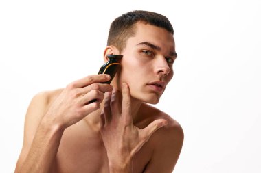 Beyaz stüdyo geçmişine karşı traş olmak için elektrikli tıraş makinesi kullanan yakışıklı genç bir adam. Günaydın cilt bakımı. Erkek güzelliği, cilt bakımı, kaplıca, kozmetoloji, erkek sağlığı kavramı