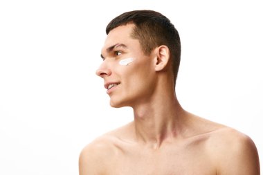 Kısa saçlı yakışıklı genç adam beyaz stüdyo geçmişine yüz nemlendirici krem sürüyor. Organik ürünler. Erkek güzelliği, cilt bakımı, kaplıca, kozmetoloji, erkek sağlığı kavramı
