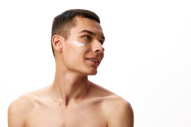 Kısa saçlı yakışıklı genç adam beyaz stüdyo geçmişine yüz nemlendirici krem sürüyor. Organik ürünler. Erkek güzelliği, cilt bakımı, kaplıca, kozmetoloji, erkek sağlığı kavramı