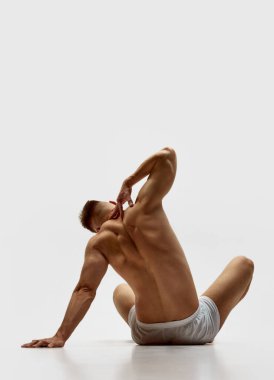 Kabarık vücutlu genç kaslı bir adam, beyaz stüdyo arka planında izole edilmiş boxerlar içinde üstsüz oturan güçlü bir sırt. Esniyorum. Erkek güzelliği, vücut bakımı, spor, sağlık