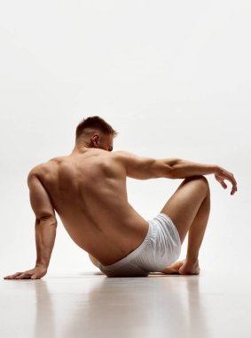 Kabarık vücutlu genç kaslı bir adam, beyaz stüdyo arka planında izole edilmiş boxerlar içinde üstsüz oturan güçlü bir sırt. Erkek güzelliği, vücut bakımı, spor, sağlık