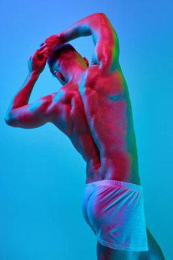 Üstsüz erkek modelin arkadan görünüşü, sırtı güçlü, kaslı vücudu pantolonun içinde, neon ışıklarındaki mavi renk stüdyo arka planına karşı duruyor. Güzellik kavramı, vücut bakımı, spor, sağlık