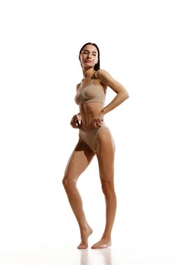 İnce vücutlu, bej rengi pamuklu iç çamaşırlarıyla beyaz stüdyo arka planında duran genç bir kadın resmi. Kadın güzelliği, vücut ve cilt bakımı, kozmetoloji, spor, sağlık