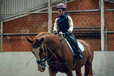 Küçük bir kız, üniformalı ve kasklı bir çocuk at üstünde oturuyor, yağmur yağıyor, özel arenada ata binmeyi öğreniyor, köşkte. Spor anlayışı, çocukluk, okul, kurs, aktif yaşam tarzı, hobi