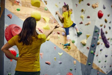Genç bir kadın, kaya terbiyecisi küçük bir kıza kaya hareketlerini, teknikleri öğretiyor. Küçük kız duvarlara tırmanıyor, kapalı alan sınıfında. Spor tırmanışı, hobi, aktif yaşam tarzı, okul, kurs kavramı