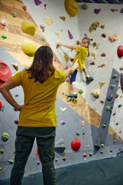 Kadın kaya terbiyecisi küçük bir kıza eğitim veriyor, çocuk kaya yapma aktivitesi, teknikler. Küçük kız duvarlara tırmanıyor, kapalı alan sınıfında. Spor tırmanışı, hobi, aktif yaşam tarzı, okul, kurs kavramı