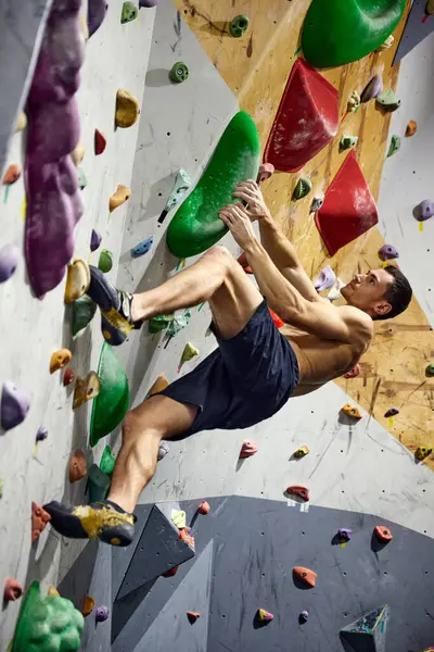 Shirtless, muscular young man climbing artificial rocks on climbing wall, practicing bouldering activity indoors. Concept of sport climbing, hobby, active lifestyle, school, training course