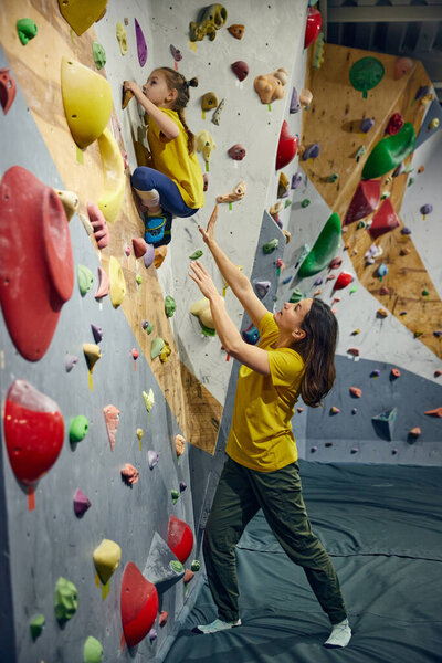 Woman, bouldering trainer teaching child little girl bouldering activity, techniques. Little girl climbing wall, indoor class. Concept of sport climbing, hobby, active lifestyle, school