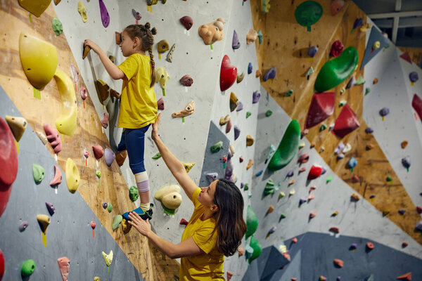Young woman, bouldering trainer teaching little girl bouldering activity, techniques. Little girl climbing wall, indoor class. Concept of sport climbing, hobby, active lifestyle, school, course