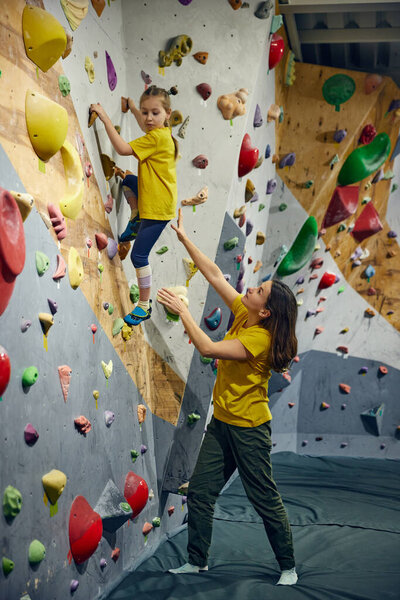 Woman, bouldering trainer teaching child little girl bouldering activity, techniques. Little girl climbing wall, indoor class. Concept of sport climbing, hobby, active lifestyle, school
