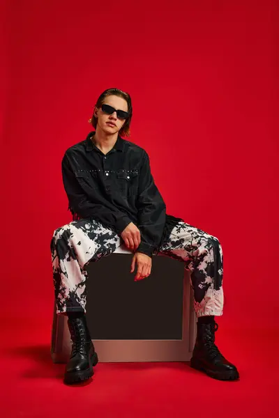 Young man, dressed as rock star in old fashioned outfit and sitting on retro TV with blank screen against red vibrant background. Concept of hobby, technology and people, retro, vintage, fashion. Ad