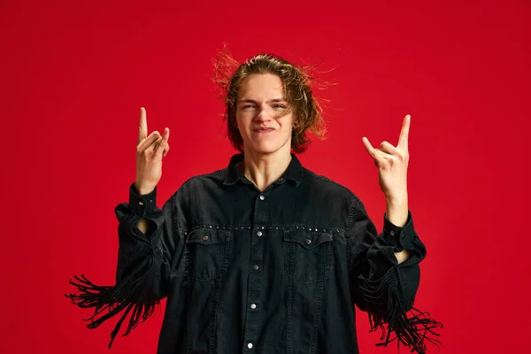 Portrait of cool attitude, young handsome man, dancing on music and showing rock-n-roll gesture against red studio background. Concept of hobby, music, retro, vintage, fashion and style. Ad