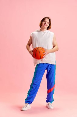 Retro spor üniforması giymiş ve basketbol topunu pastel pembe arka planda tutan genç bir adamın tam boy portresi. Aktif yaşam tarzı, spor ve eğlence anlayışı, hobi, moda.