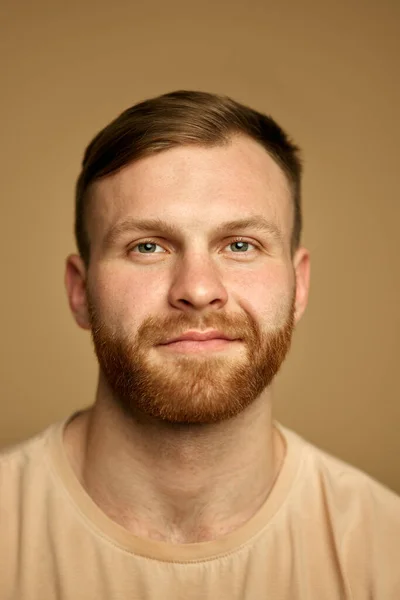 ハンサムな赤毛の若い男の垂直な肖像画は 砂色のスタジオの背景に対してカメラを見ているひげを微笑みます セルフケア 化粧品 スパのコンセプト — ストック写真