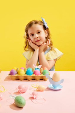Güzel küçük bir kızın portresi, sarı arka planda paskalya yumurtalarıyla masada oturan yay tüylü aksesuarlı bir çocuk. Paskalya tatili kavramı, kutlama, gelenekler, çocukluk, mutluluk