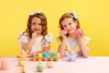 Şirin küçük kızlar, sarı arka planda boyalı Paskalya yumurtalarıyla masada oturan elbiseli çocuklar. Geleneklere uymak. Paskalya tatili, kutlama, çocukluk, mutluluk kavramı