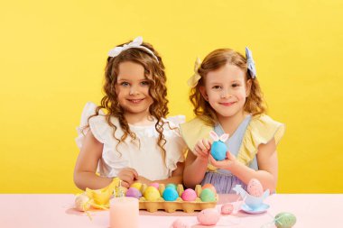 Küçük kızlar, güzel elbiseli çocuklar masa boyama ve sarı arka planda Paskalya yumurtaları süsleme. Paskalya tatili kavramı, kutlama, gelenekler, çocukluk, mutluluk