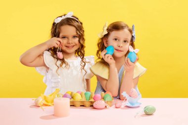 Güzel küçük kızlar, ellerinde boyalı elbiseler ve sarı arka planda süslü Paskalya yumurtaları olan kız kardeşler. Paskalya tatili kavramı, kutlama, gelenekler, çocukluk, mutluluk