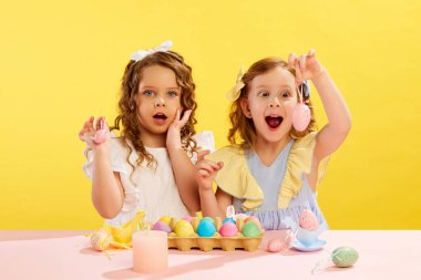 Duygusal küçük kızlar, çocukların yaptıkları, sarı arka planda Paskalya yumurtaları süslemeleri ve boyamaları. Paskalya tatili kavramı, kutlama, gelenekler, çocukluk, mutluluk