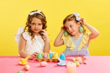 Güzel küçük kızların portresi, masada oturan çocuklar, sarı arka planda boyanmış yumurtalar. Paskalya tatili kavramı, kutlama, gelenekler, çocukluk, mutluluk