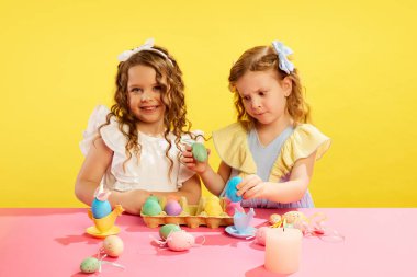 Kız kardeşiyle oturan küçük kız, tuhaf bir şekilde sarı arka plandaki Paskalya yumurtalarına bakıyor. Renkli yumurta resimleri. Paskalya tatili kavramı, kutlama, gelenekler, çocukluk, mutluluk