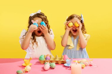 Duygusal, heyecanlı, gözleri boyalı Paskalya yumurtaları tutan küçük kızlar, sarı arka planda, masada duruyorlar. Paskalya tatili kavramı, kutlama, gelenekler, çocukluk, mutluluk