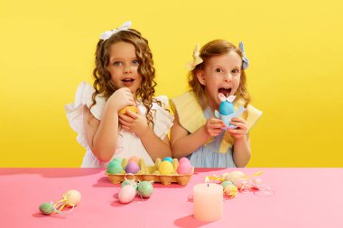 Sarı arka planda boyalı Paskalya yumurtalarıyla dikilen güzel elbiseli duygusal komik küçük kızlar. Paskalya tatili kavramı, kutlama, gelenekler, çocukluk, mutluluk