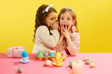 Küçük bir kız kulağına fısıldıyor, çocuklar mutlu ve heyecanlı bir yüz ve sarı bir arka plan. Paskalya tatili kavramı, kutlama, gelenekler, çocukluk, mutluluk