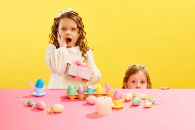 Duyarlı küçük çocuklar hediyelerle ayakta duruyor, boyalı ve süslü Paskalya yumurtalarını sarı arka plana dikizliyorlar. Paskalya tatili kavramı, kutlama, gelenekler, çocukluk, mutluluk