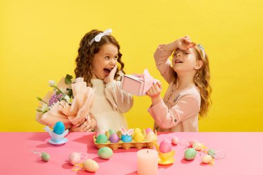 Küçük kızlar Paskalya bayramını kutluyorlar, ellerinde hediye kutuları, çiçekler, sarı arka planda boyalı yumurtalarla masada duruyorlar. Paskalya tatili kavramı, kutlama, gelenekler, çocukluk, mutluluk
