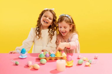 Küçük kızlar, çocuklar neşeyle gülüyor, gülümsüyor, sarı arka planda süslü ve boyalı yumurtalarla masada oturuyorlar. Paskalya tatili kavramı, kutlama, gelenekler, çocukluk, mutluluk