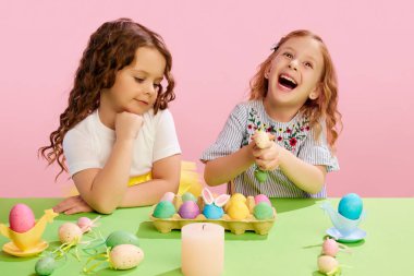 Mutlu küçük kızlar dekore ediyor, Paskalya yumurtaları boyuyor, gülümsüyor ve pembe arka planda eğleniyorlar. Paskalya tatili kavramı, kutlama, gelenekler, çocukluk, mutluluk