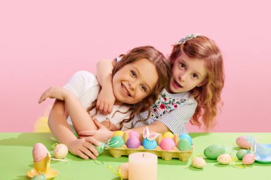 Masada süslü, boyalı yumurtalarla oturan, sarılan ve pembe arka planda gülümseyen güzel kızların portresi. Paskalya tatili kavramı, kutlama, gelenekler, çocukluk, mutluluk