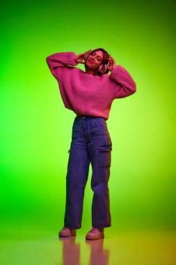 Kulaklıkla müzik dinleyen aşırı neşeli genç bir kadının tam boy portresi pembe neon ışıkta yeşil gradyan arka planda. Müzik ve dans kavramı, parti, teknoloji, kültür, hareket. Ad