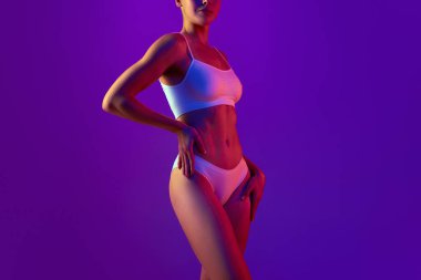 Spor ve diyet programı. İnce, kaslı, beyaz iç çamaşırlı atletik kadın vücudunun kesilmiş görüntüsü neon ışıkta mor arka planda. Doğal güzellik kavramı, sağlık ve vücut bakımı, spor